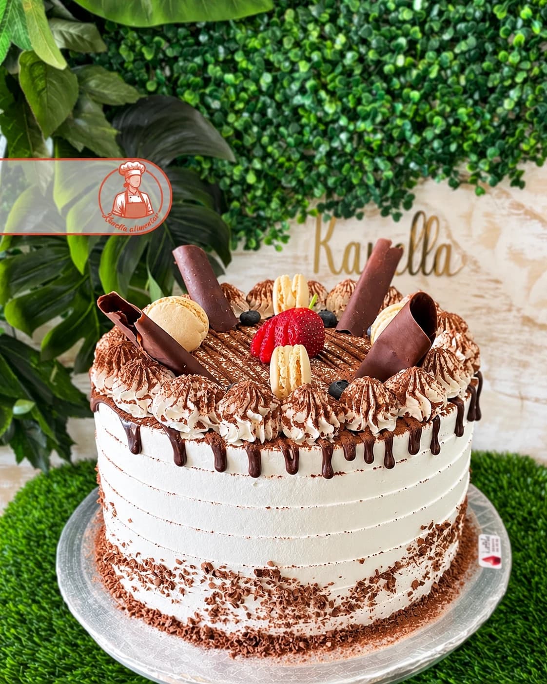 Bolo com creme branco, cacau, cachos de chocolate, macarons e morango, junto ao letreiro dourado Kanella