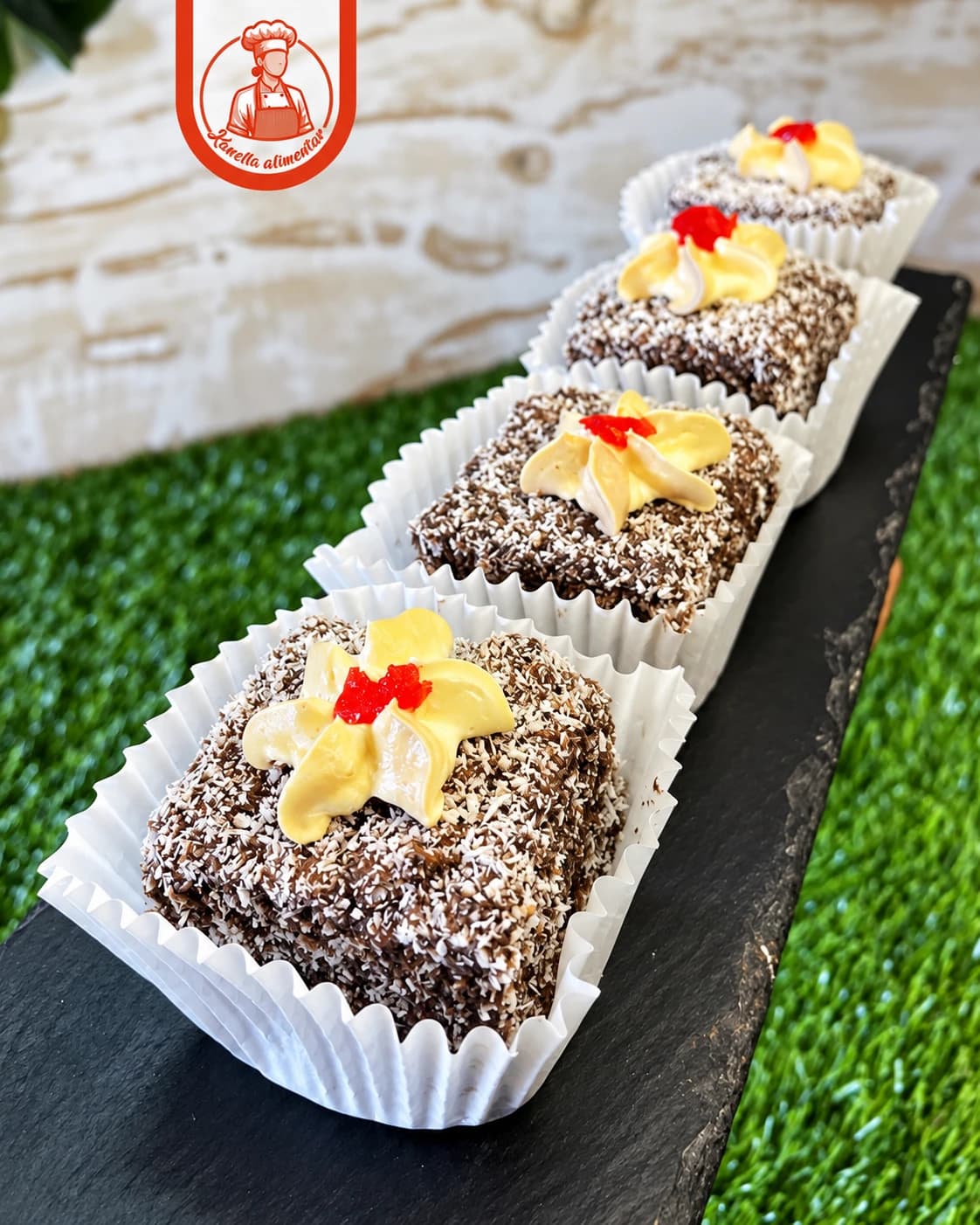 Quadrados de chocolate e coco com creme amarelo e cereja, em forminhas brancas sobre uma ardósia
